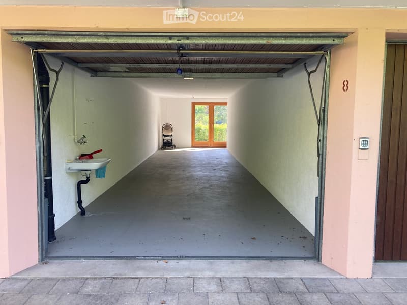 A narrow, concrete-floored corridor leading to a garage area with a sink and a stroller visible at the end.
