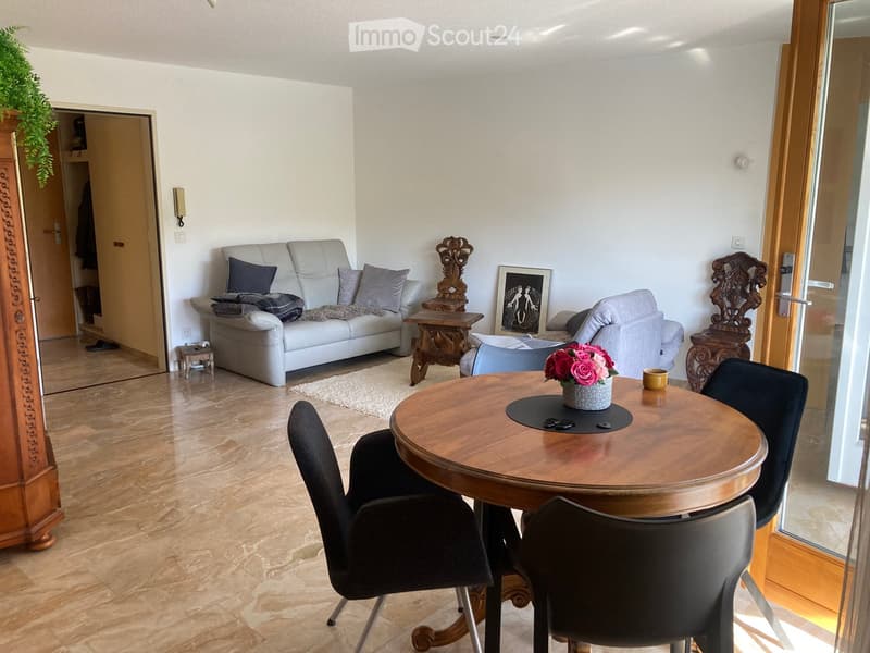Bright living room with a white sofa, armchair, coffee table, dining table, and chairs. Wooden flooring, large windows, and decorative items.