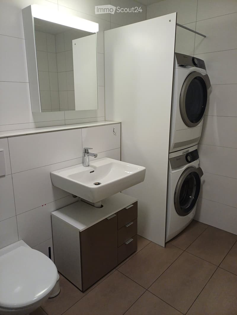 Modern bathroom with a white sink, mirror, cabinet, toilet, and a stacked washing machine and dryer unit.
