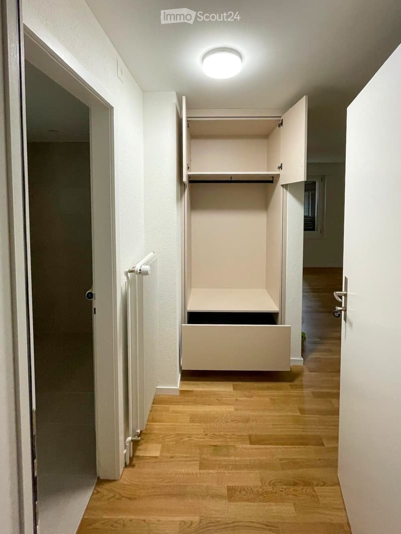 A narrow hallway with wooden flooring, white walls, and a built-in closet with open shelves and drawers. The hallway features a ceiling light and a radiator on the left wall.