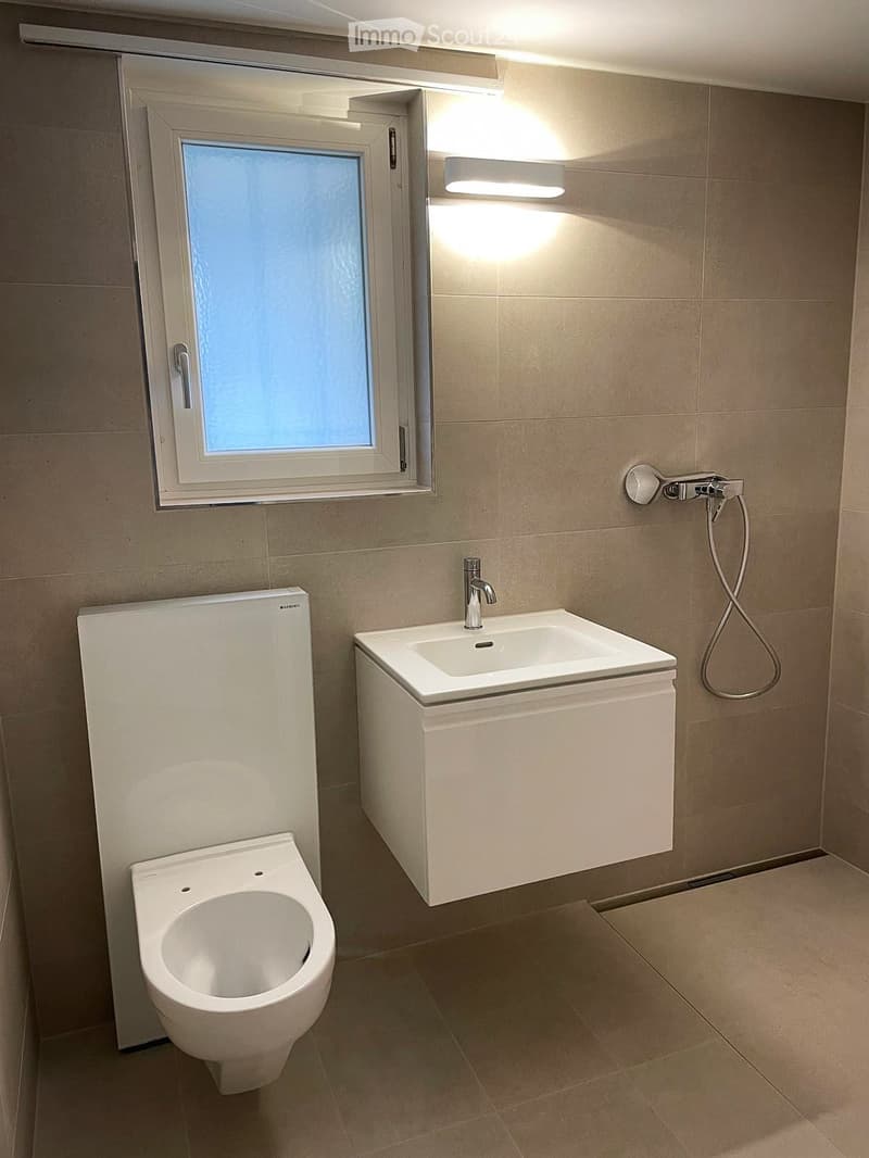 Modern bathroom with a white sink, toilet, and showerhead, featuring beige tiled walls and floor, and a window with frosted glass.