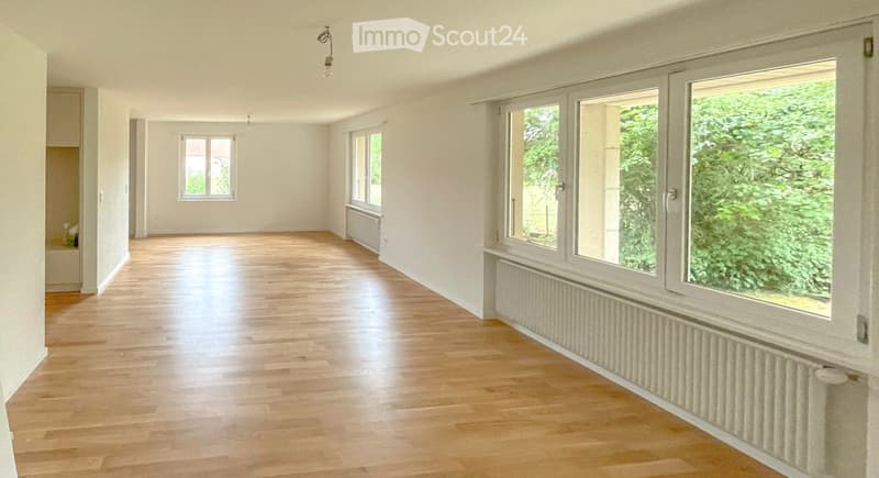 Empty room with wooden flooring, white walls, and large windows offering a view of greenery outside. No furniture or equipment is present.