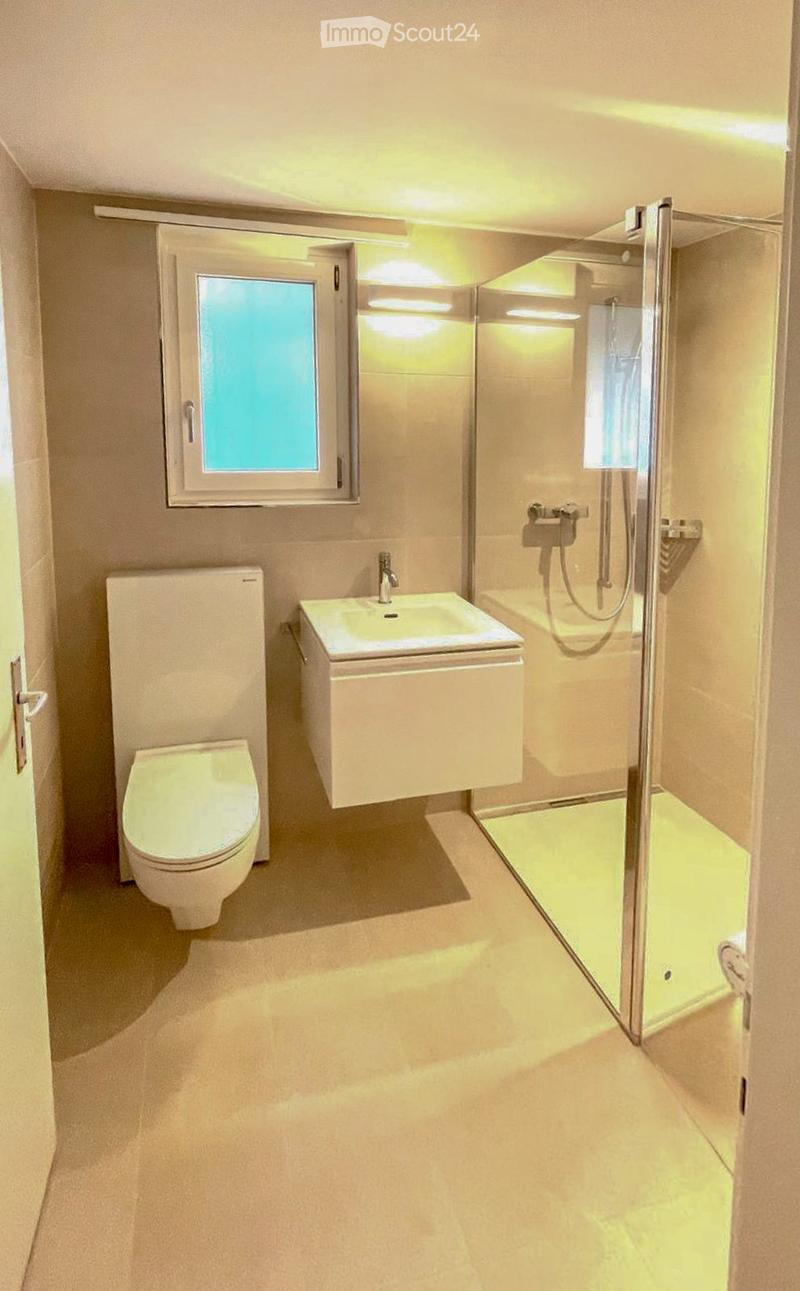 Modern bathroom with a toilet, sink, and glass-enclosed shower stall. Features a window with frosted glass and built-in lighting.