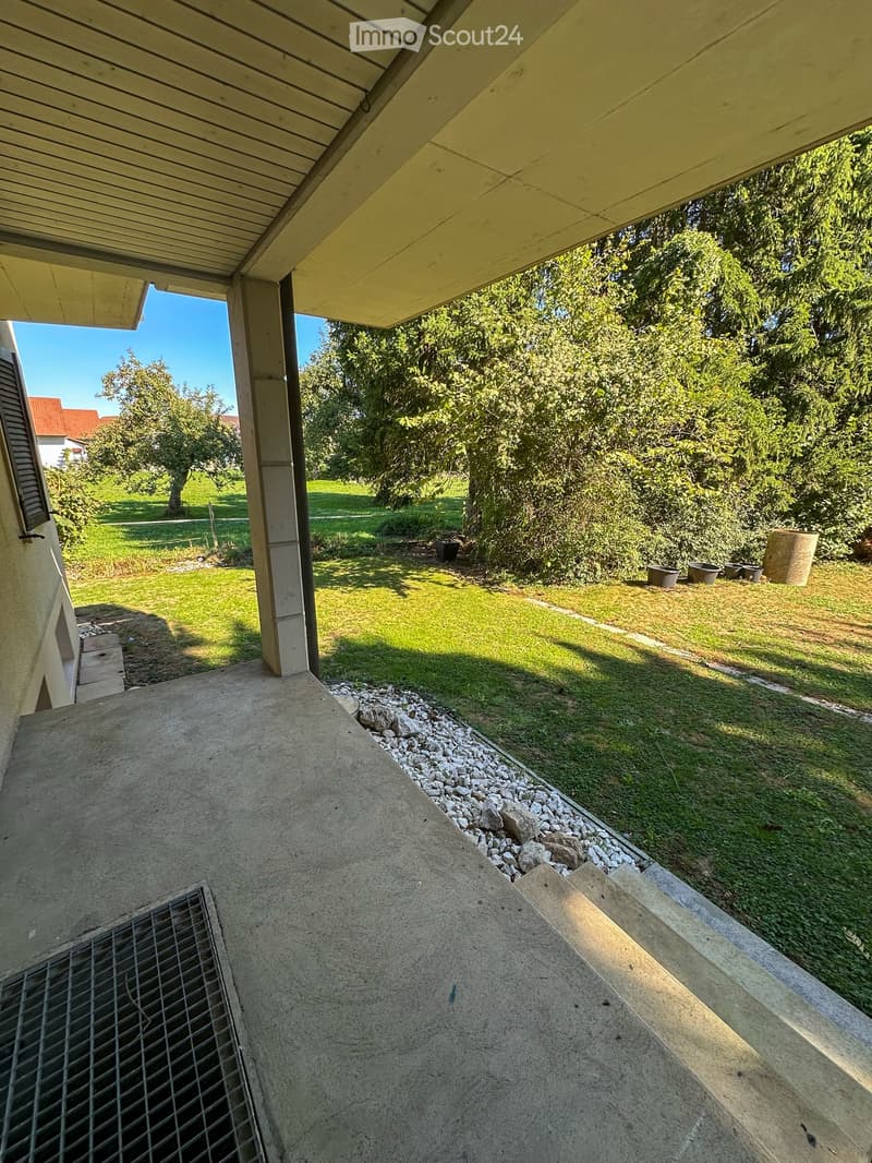 A covered patio area with a concrete floor, surrounded by a well-maintained lawn and trees. The area includes a metal grate and a small garden bed with rocks and potted plants.