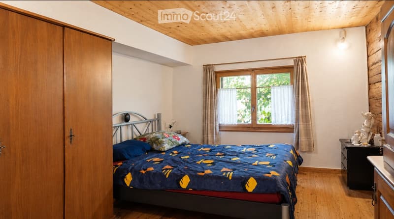 Cozy bedroom with wooden ceiling, wooden floor, large window with curtains, bed with colorful bedding, nightstand with mirror, and decorative statue on the nightstand.