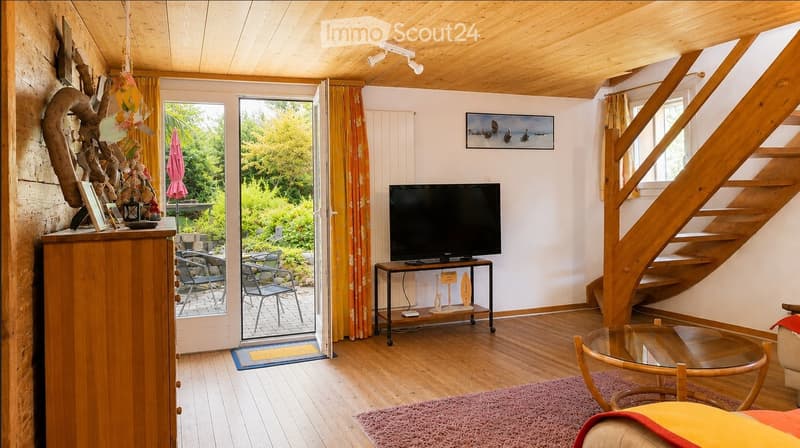 A cozy living room with wooden floors, a television on a stand, a glass coffee table, and a staircase leading to another level. The room features a large window with a view of a garden, a wooden cabinet with decorative items, and a staircase leading to an