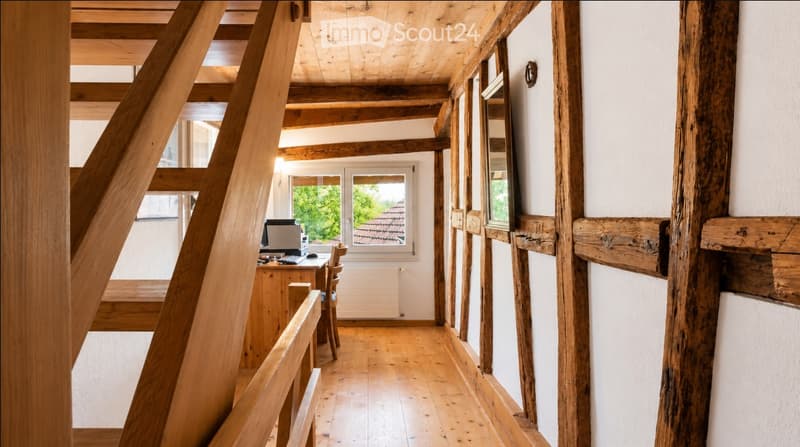 A wooden-framed room with exposed beams and a wooden floor, featuring a desk with a computer, chair, and a window overlooking a garden.