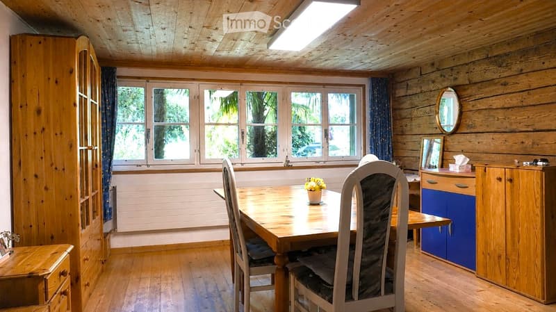 Cozy wooden-paneled room with a dining table, chairs, a cabinet, and a mirror. Large windows offer a view of greenery outside.