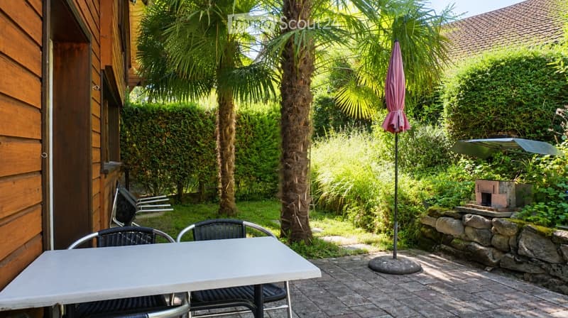 A sunny outdoor patio area with a white table, black chairs, a closed umbrella, and a small stone barbecue surrounded by lush greenery and palm trees.