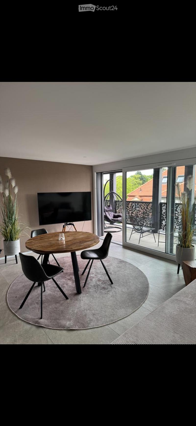 Modern dining area with a round wooden table, black chairs, flat-screen TV, and large glass sliding doors leading to a patio with outdoor seating and plants.