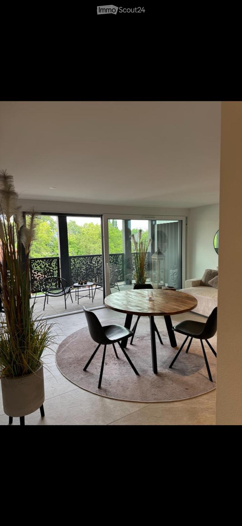 Modern dining area with a round wooden table, black chairs, a sofa, and a small side table, connected to a balcony with outdoor seating.