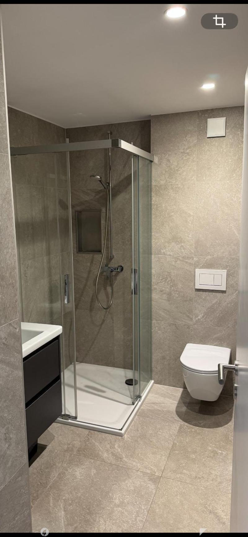 Modern bathroom with a glass-enclosed shower, toilet, and sink. The walls and floor are tiled in a neutral tone, and the shower features a handheld sprayer.