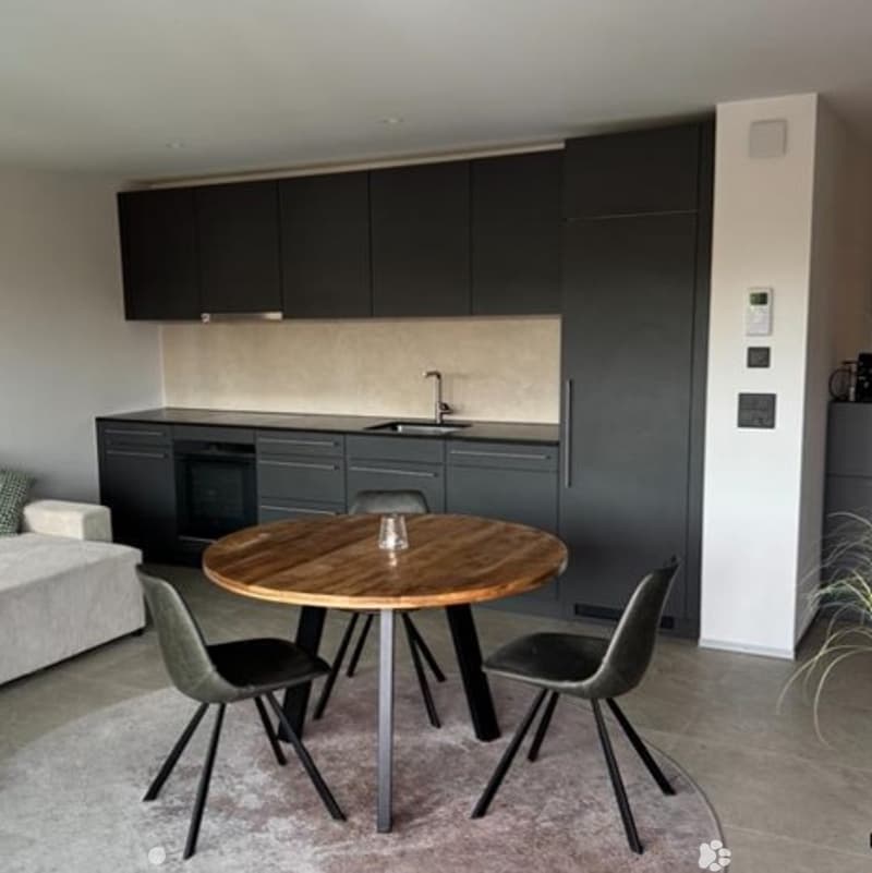 Modern kitchen with dark grey cabinets, a sink, and an oven. A round wooden dining table with four grey chairs is placed in the foreground, and a white sofa is visible to the left.