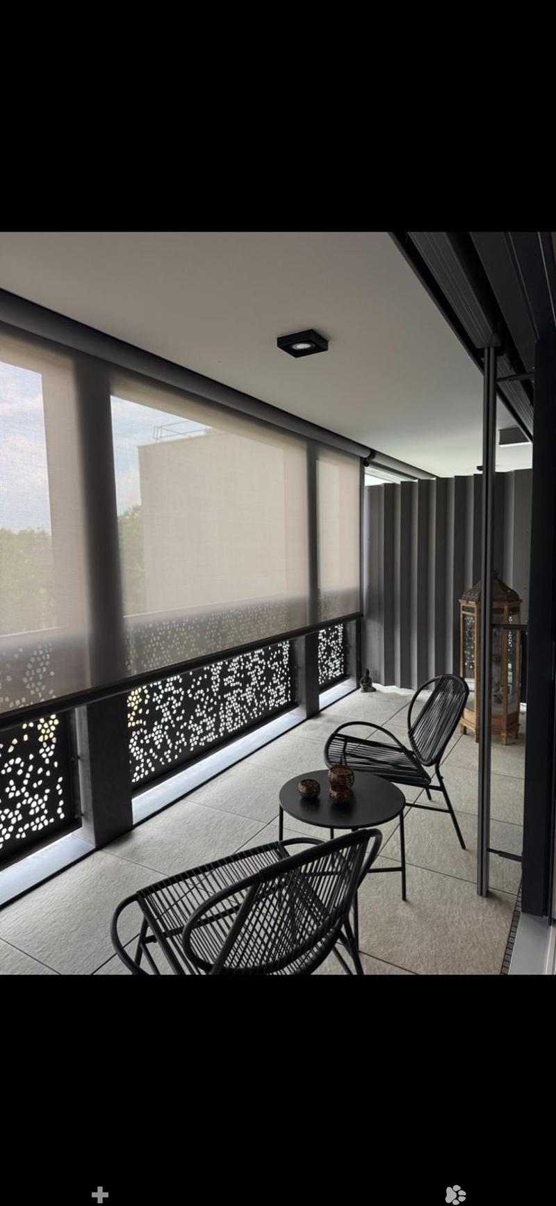 Modern balcony with large windows, black metal chairs, small round table, decorative lantern, and patterned balustrade.