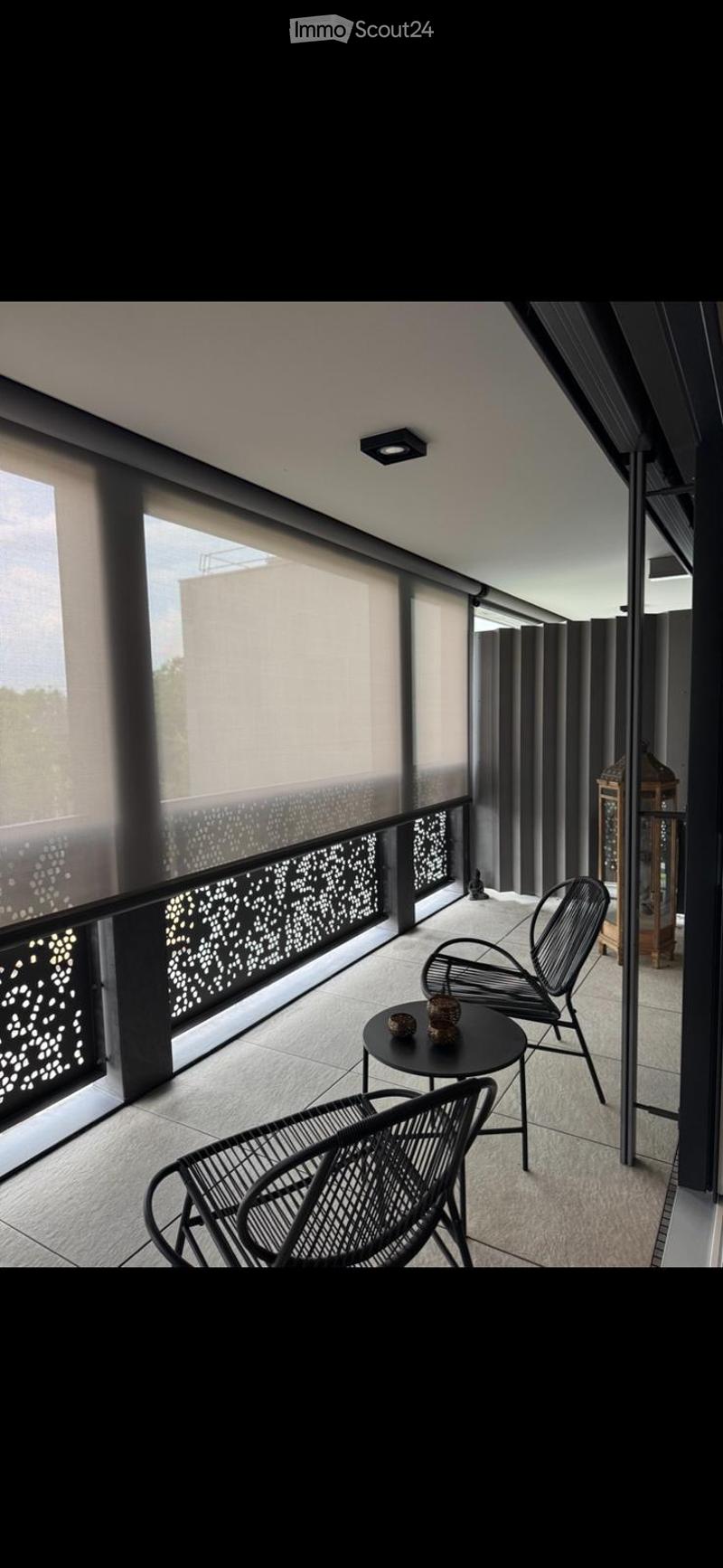Modern balcony with large windows, black metal chairs, small round table, decorative lantern, and patterned balustrade.