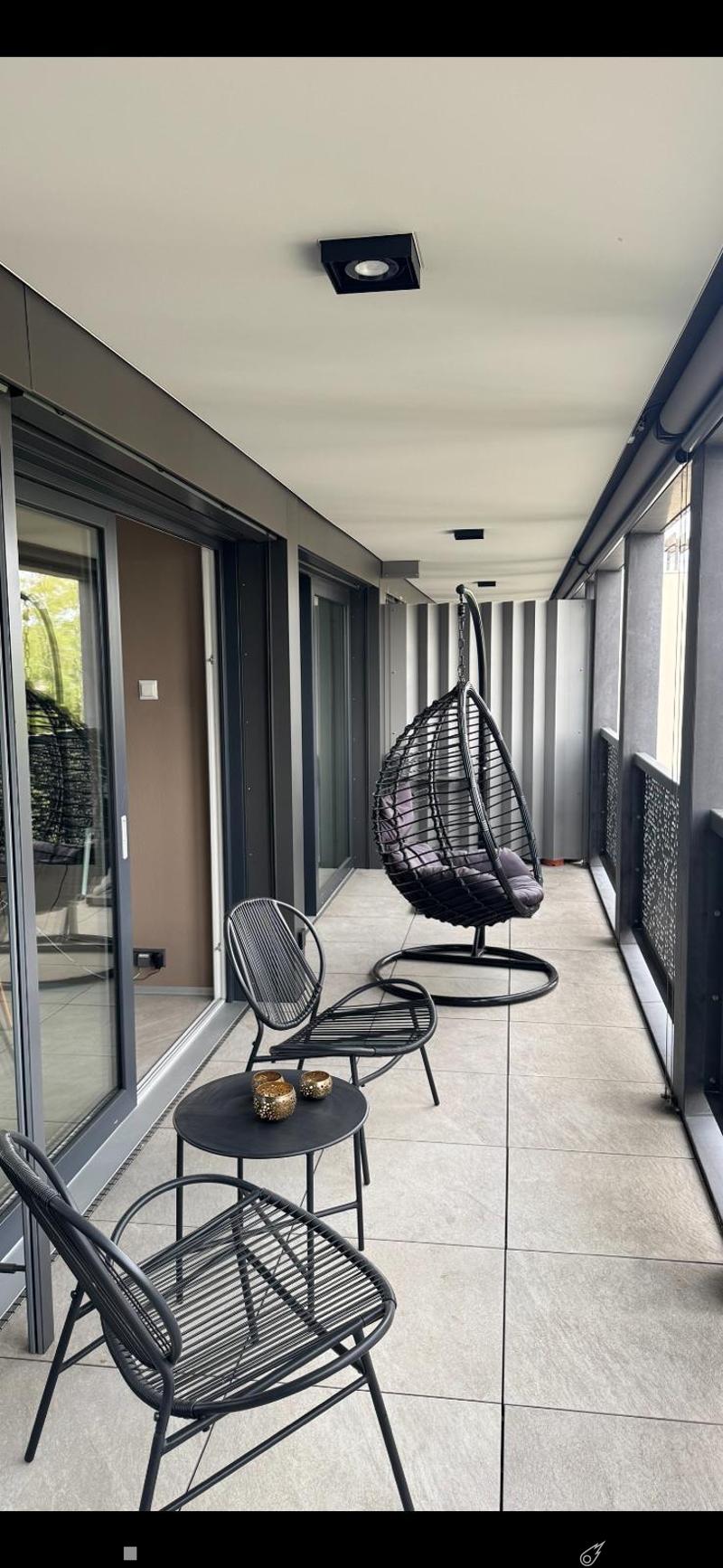 Modern balcony with tiled floor, black metal furniture including a hanging wicker chair, two metal chairs, a small table, and decorative candle holders.