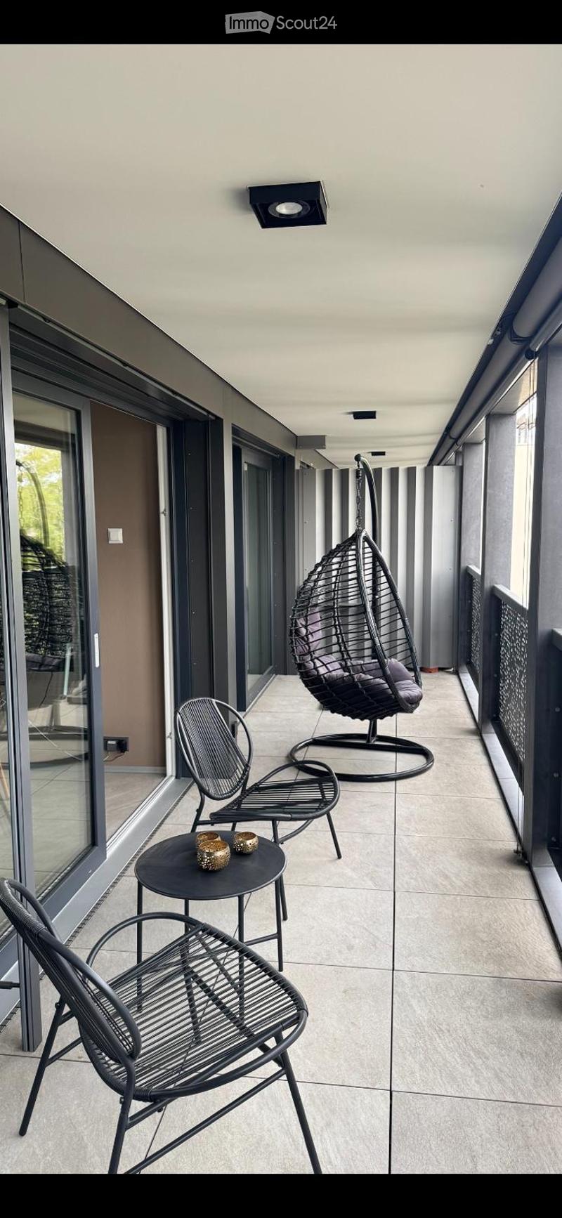 Modern balcony with tiled floor, black metal furniture including a hanging wicker chair, two metal chairs, a small table, and decorative candle holders.