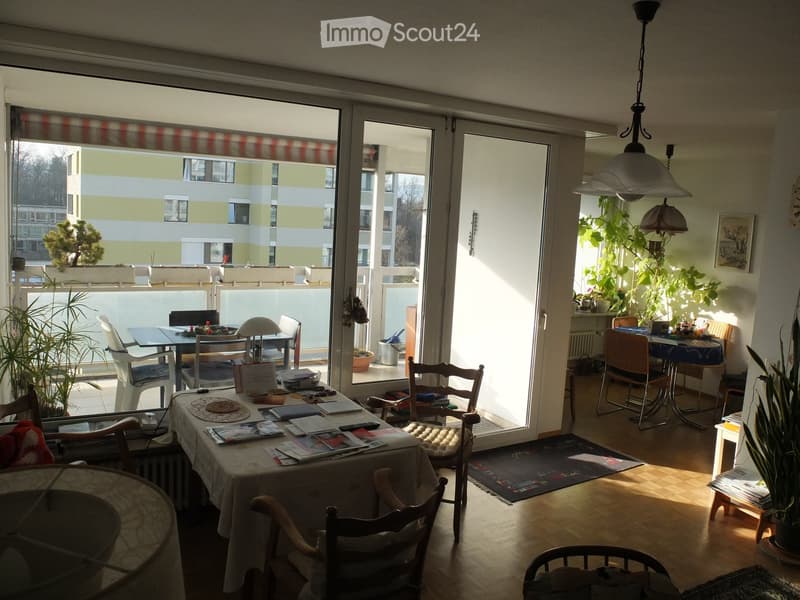 Bright living room with large windows and a balcony, featuring a dining table, chairs, a lamp, and plants.