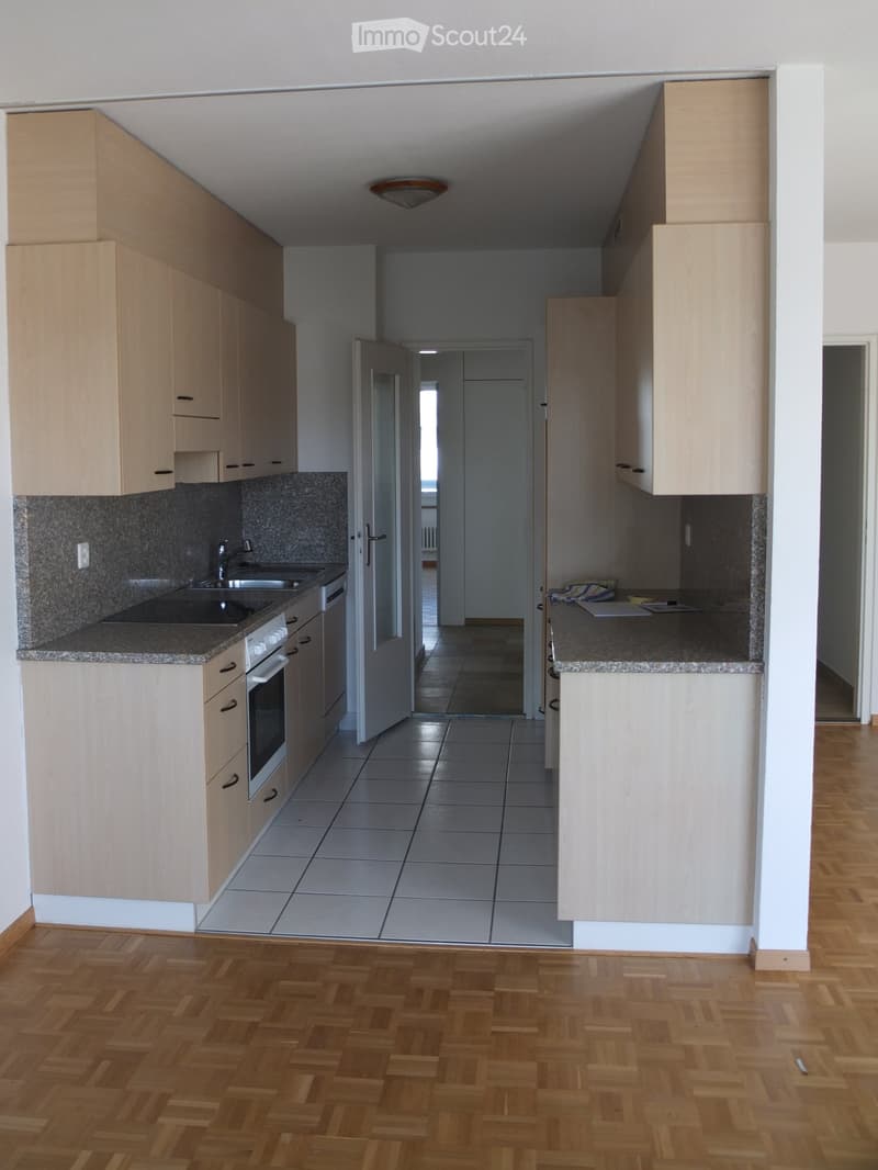 Modern kitchen with light wooden cabinets, granite countertops, and a tiled floor. The kitchen features a sink, oven, and built-in cabinets. The adjacent area shows a hallway with wooden flooring.