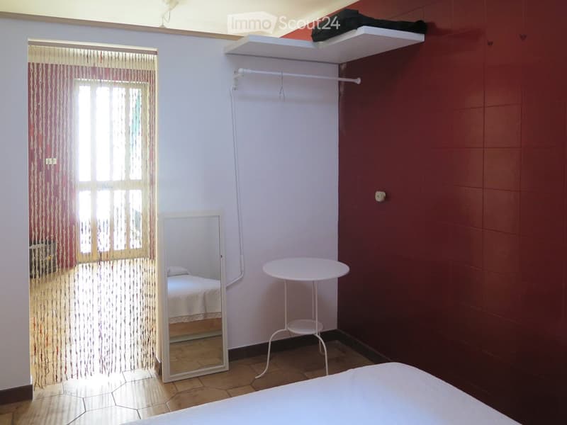 A small bedroom with a bed, a round table, a mirror, a shelf, and a clothing rack. The room has a window with a curtain and a tiled floor.