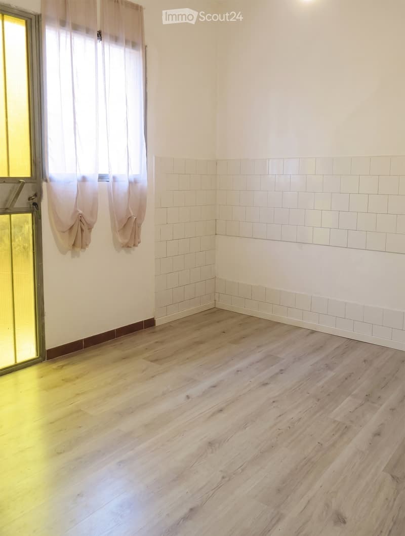 Empty room with light wooden flooring, white tiled lower walls, and a window with beige curtains. The walls are painted white, and the room has a glass door on one side.