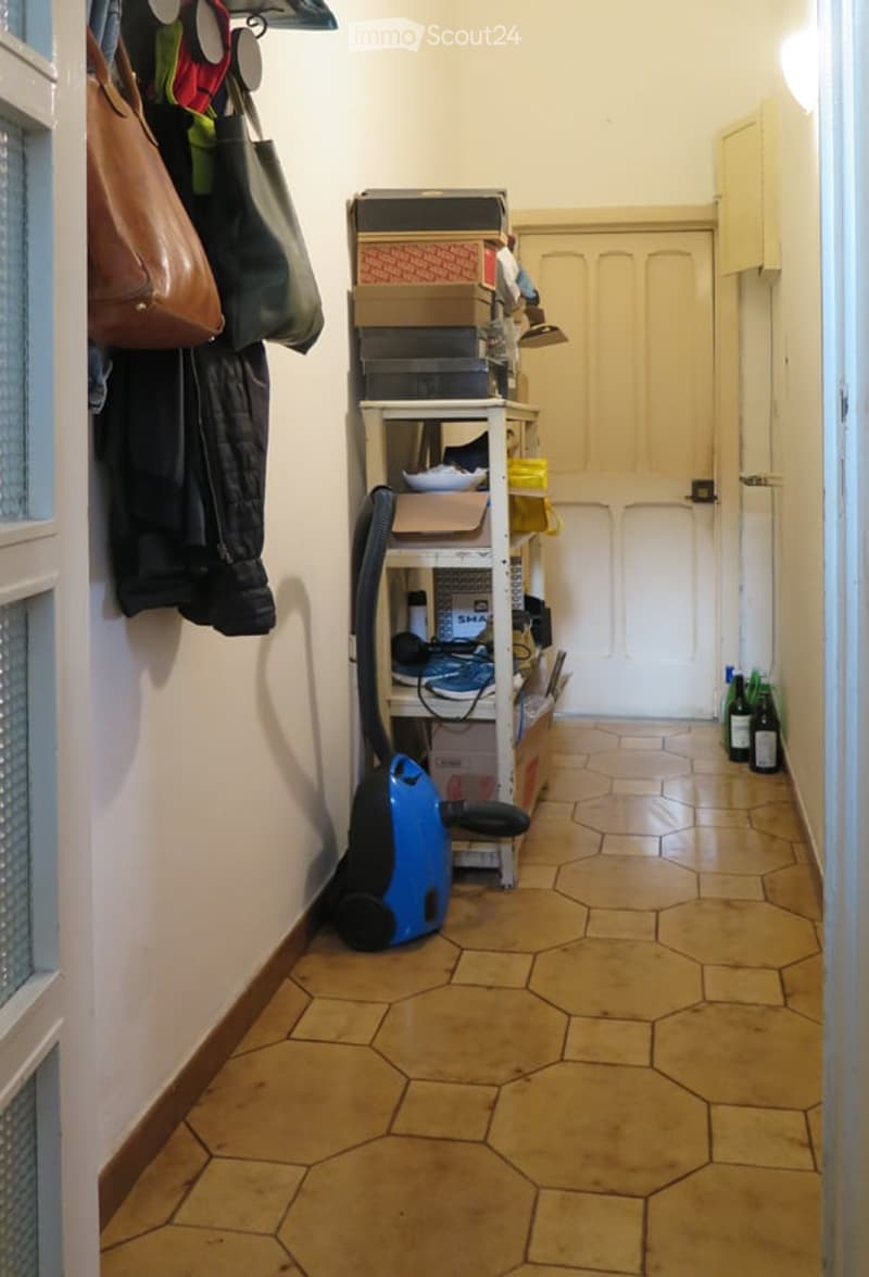 Narrow hallway with tiled floor, storage shelves, vacuum cleaner, coats, and bags hanging on the wall.