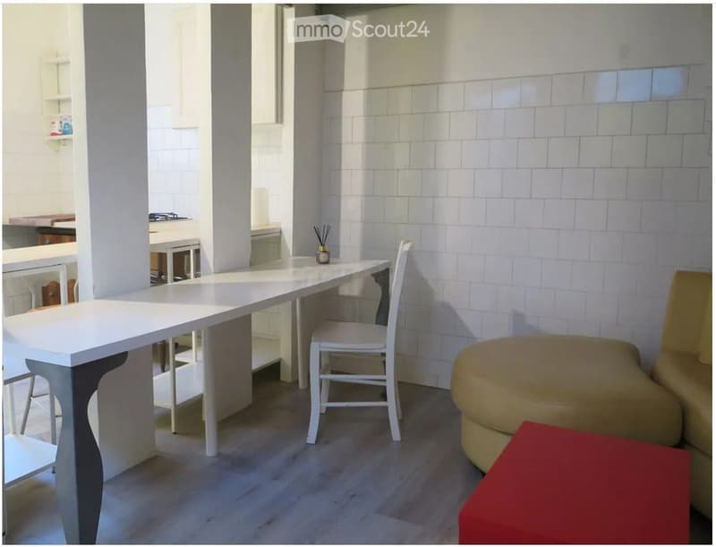 A modern, minimalist room with a white table, white chair, beige ottoman, and red square stool. The walls are white tiled, and the floor is light wood.