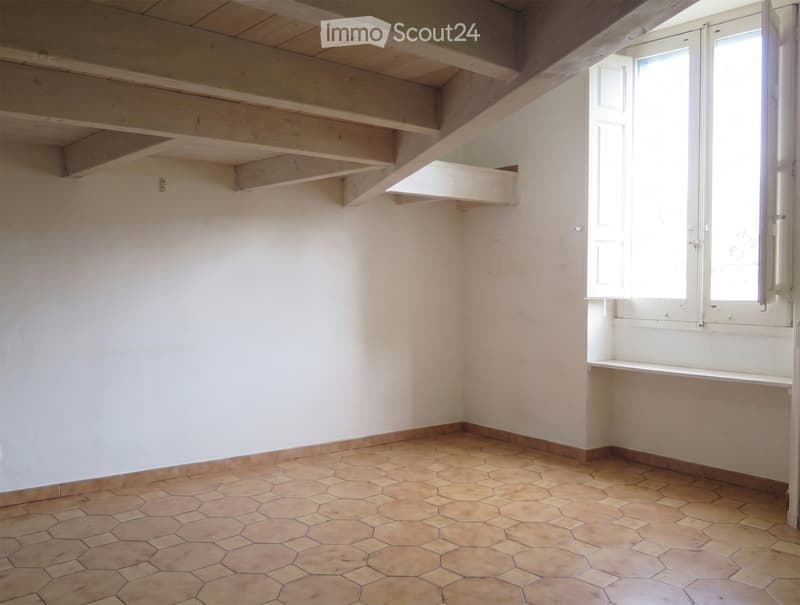 Empty room with exposed wooden beams on the ceiling, hexagonal tiled floor, and a window with shutters on the right side.