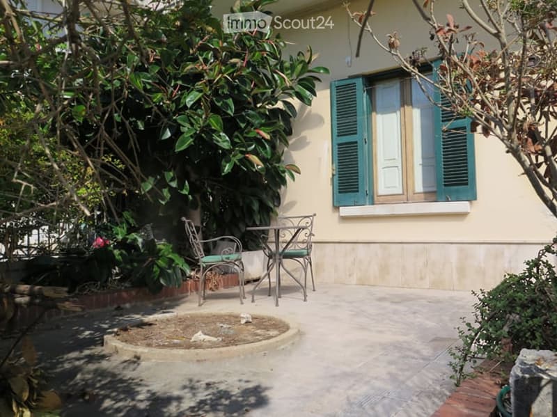 Small outdoor patio area with a round concrete planter, a metal table, and two chairs. Greenery surrounds the area, and a window with shutters is visible in the background.