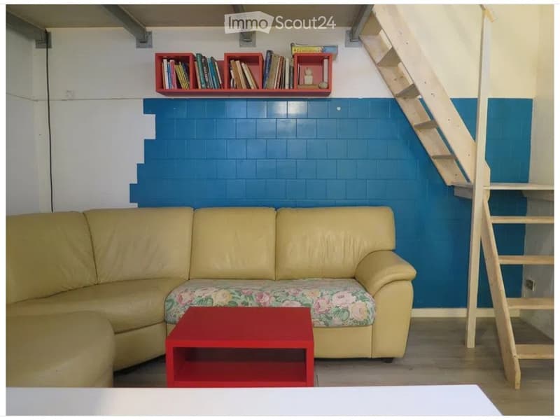 A modern living room with a cream-colored sectional sofa, a red coffee table, and a blue accent wall. A wooden staircase is visible on the right side, and a red bookshelf with books is mounted on the wall.