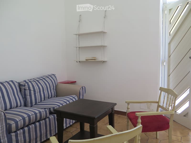 Living room with a blue and white striped sofa, a dark wooden coffee table, a white wooden chair with a red cushion, and a floating shelf with books.