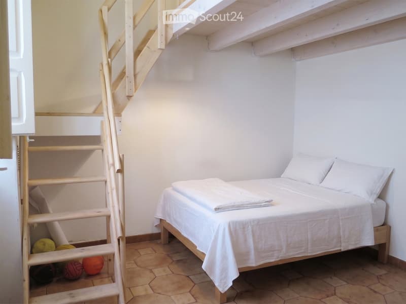 A small bedroom with a single bed, white bedding, and a wooden staircase leading to a loft area. The room has a tiled floor and white walls.