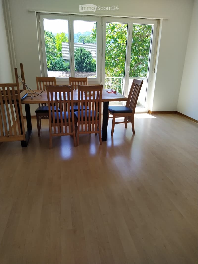 Dining room with wooden floor, dining table, and chairs, large windows with a view of greenery.