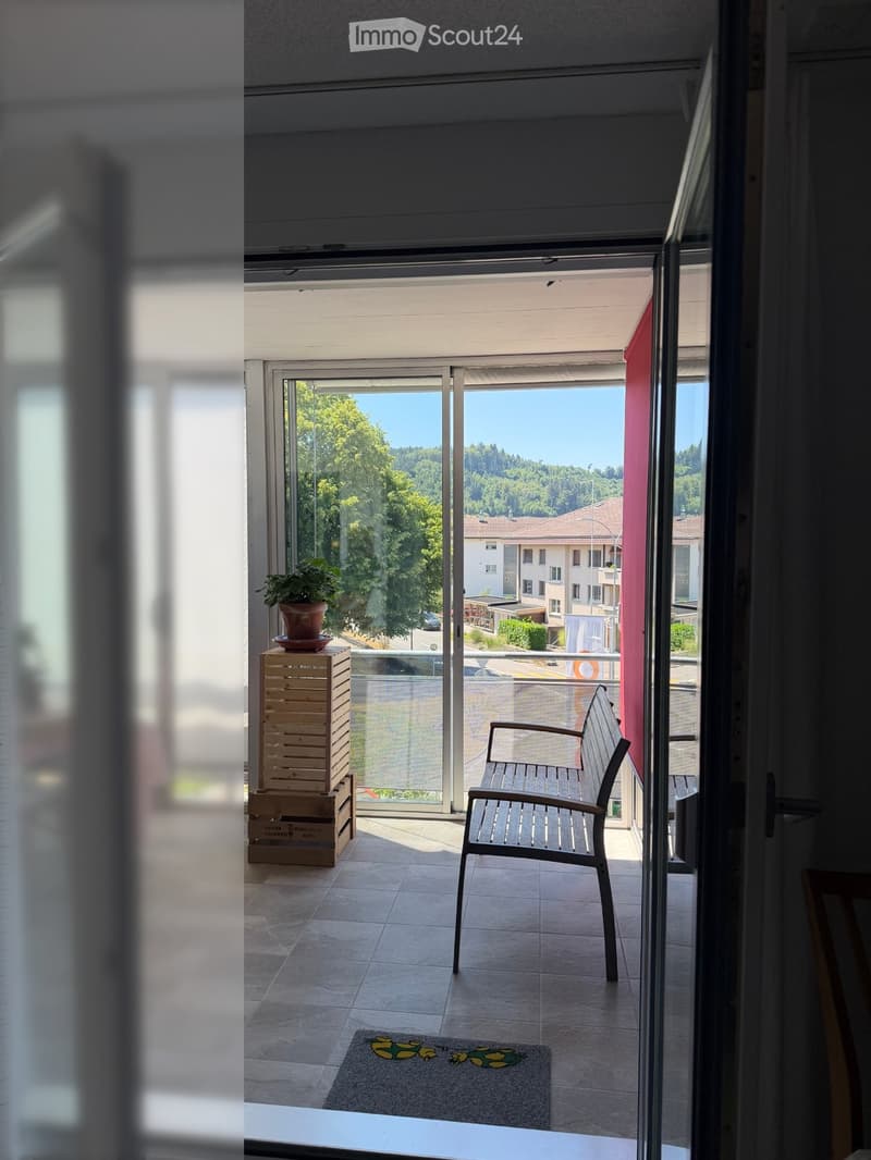 Balcony with tiled floor, wooden chair, bench, rug, and potted plant, overlooking a green landscape.