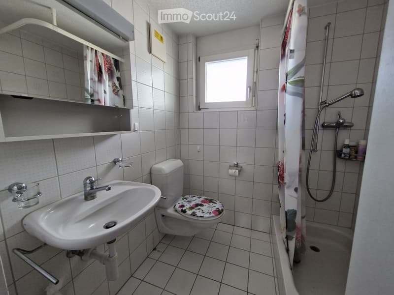 A small bathroom with a white sink, toilet, mirror, and a shower area with a curtain. The walls and floor are tiled in light gray.