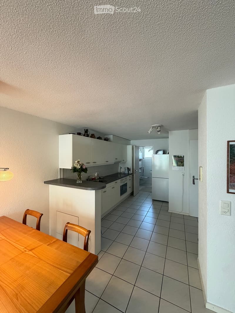 A modern kitchen with white cabinets, a wooden dining table, and a refrigerator. The space includes a sink, oven, and a vase with flowers on the counter.