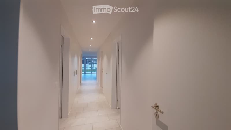 A clean, well-lit hallway with white walls, tiled flooring, and recessed ceiling lights. Doors are visible on both sides, leading to other rooms.