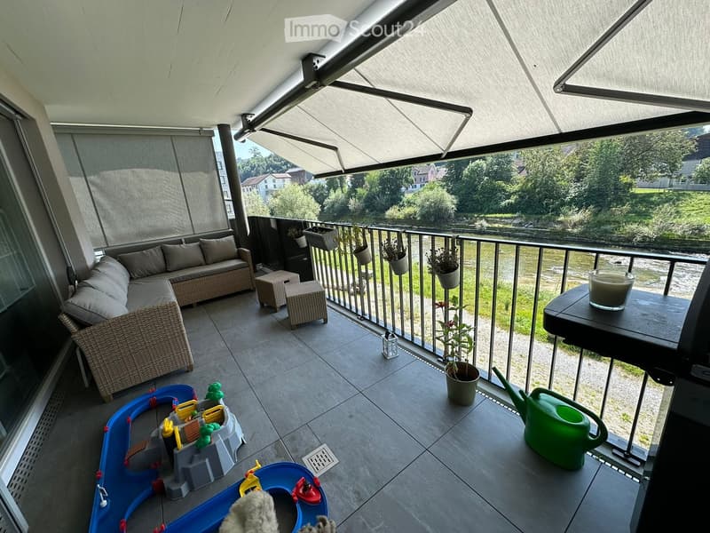 Balcony with a sofa, stools, a small table with a cup, potted plants, a toy set, and a watering can. The balcony has a retractable awning and overlooks a green landscape.