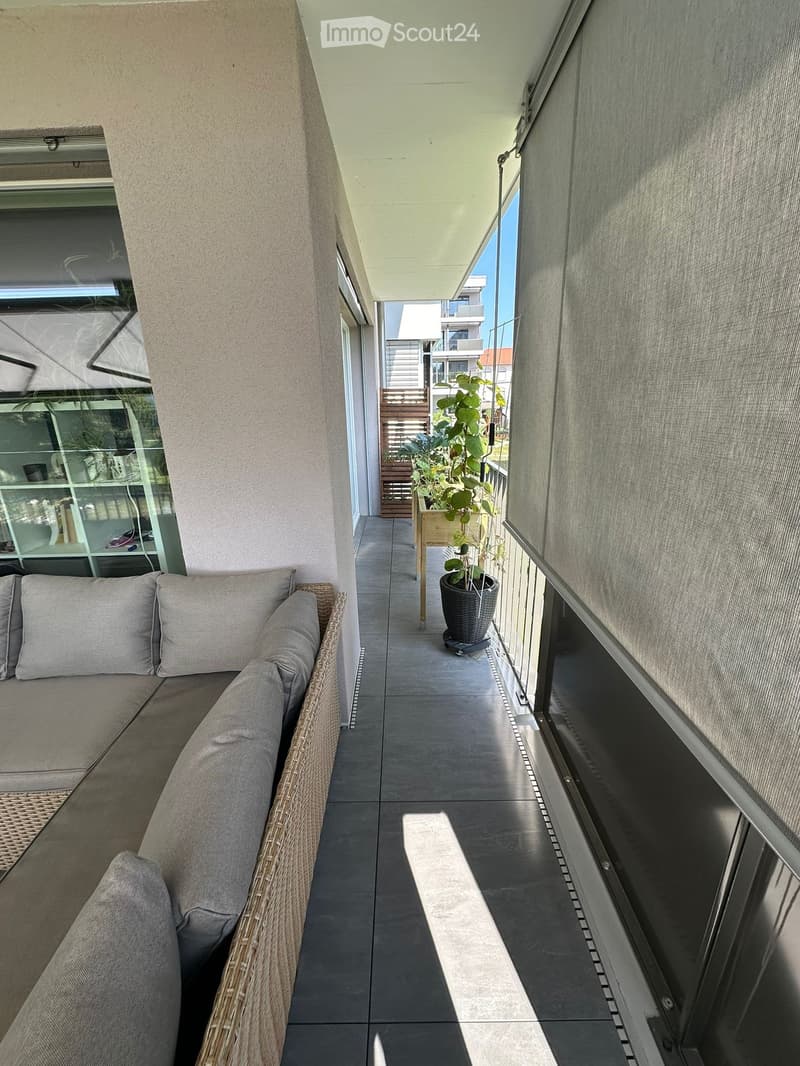 Balcony with a wicker sofa, cushions, and a potted plant, featuring a tiled floor and large windows with blinds.