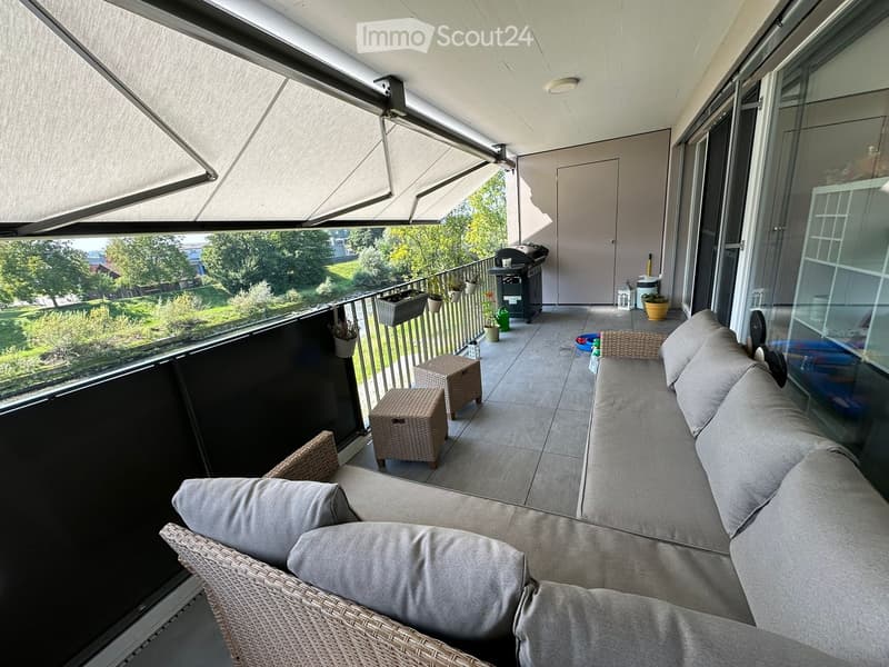 Balcony with a grey sofa, two wicker stools, a barbecue, and potted plants, featuring a retractable awning and a view of a green garden.