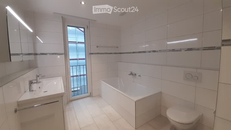 Modern bathroom with white tiles, a bathtub, sink, toilet, and a large mirror. Features a window with a view of a balcony.