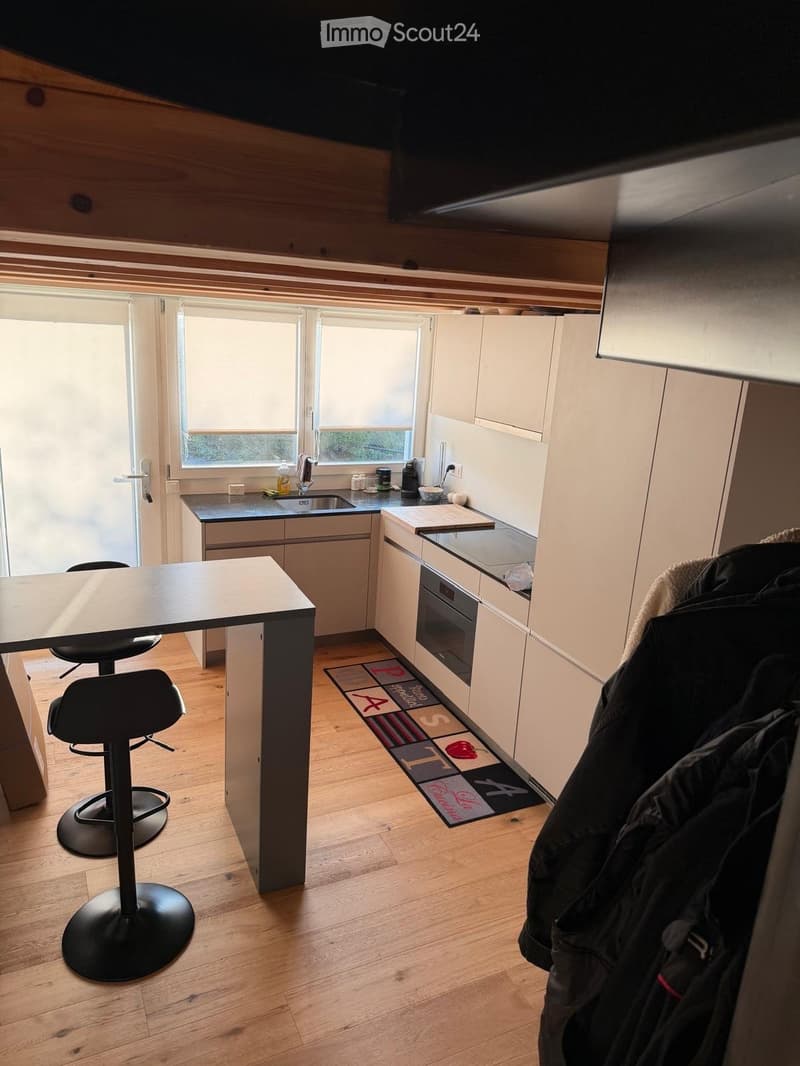 Modern kitchen with light-colored cabinets, a central island with a table and two stools, a sink, and an oven. Wooden flooring and a decorative rug are visible.
