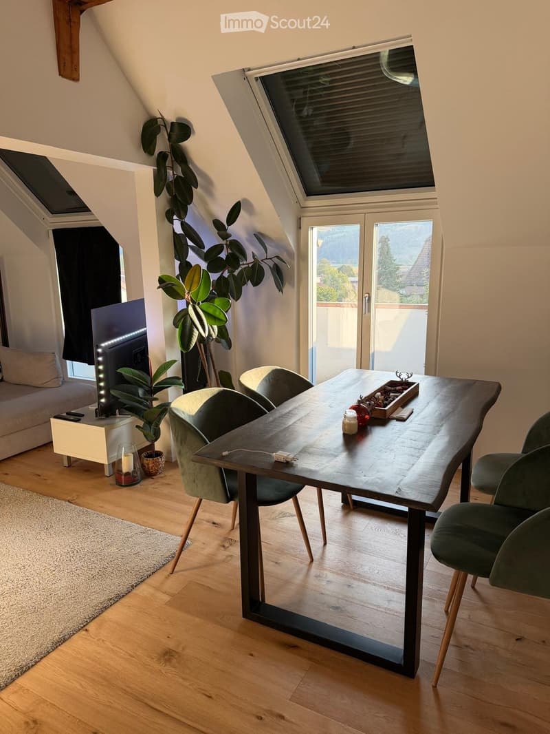 Bright living room with wooden floors, large windows, and a view. Features a dining table with chairs, a sofa, a television, and decorative plants.