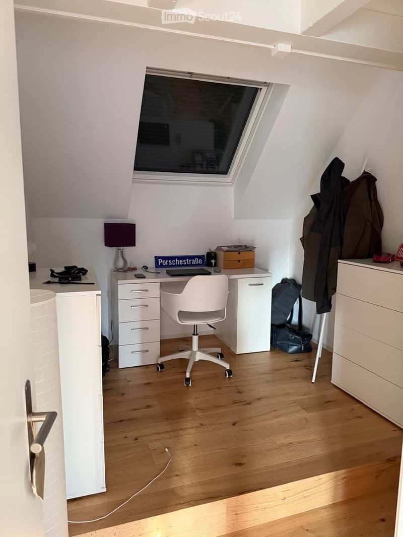 Bright, modern attic room with wooden flooring, white walls, and a large skylight window. Features a white desk with a chair, a lamp, and various items including a laptop and a backpack. A coat is hanging on the wall.