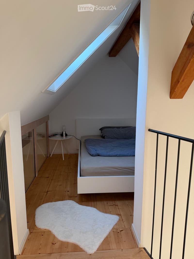 Cozy attic bedroom with wooden floors, white walls, skylight window, bed with pillows and blanket, small table with bottle, and white rug.