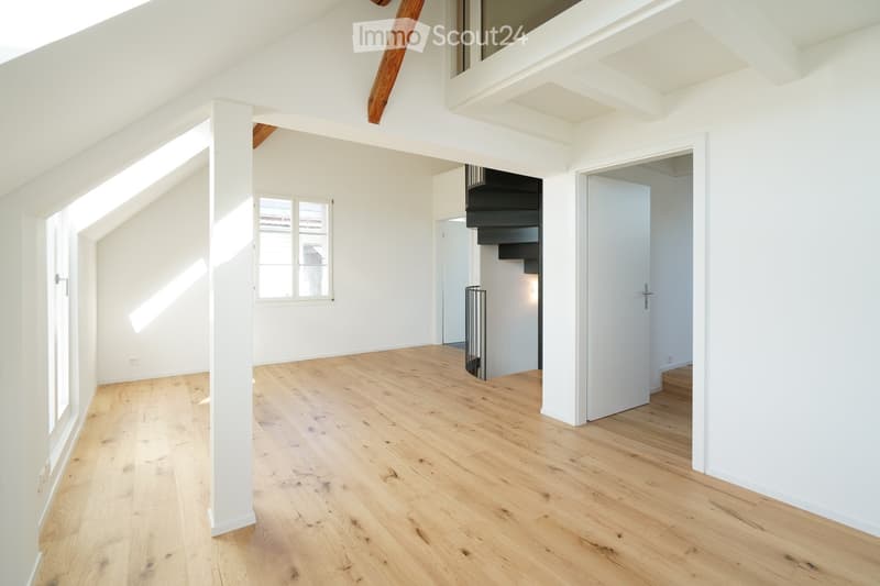 Bright, empty room with wooden flooring, white walls, and large windows allowing natural light. Features a staircase leading to another level and exposed wooden beams on the ceiling.