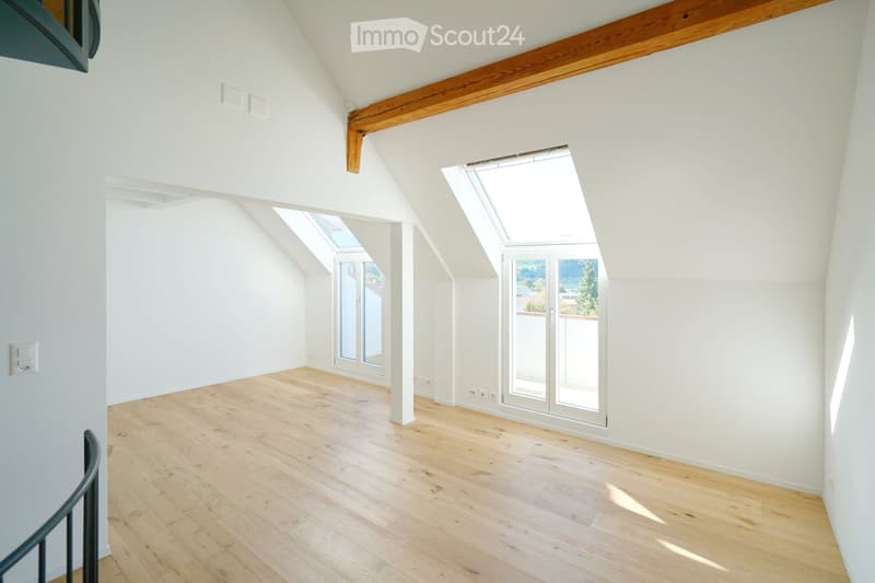Bright, empty attic room with wooden flooring, white walls, exposed wooden beams, and skylight windows offering natural light.