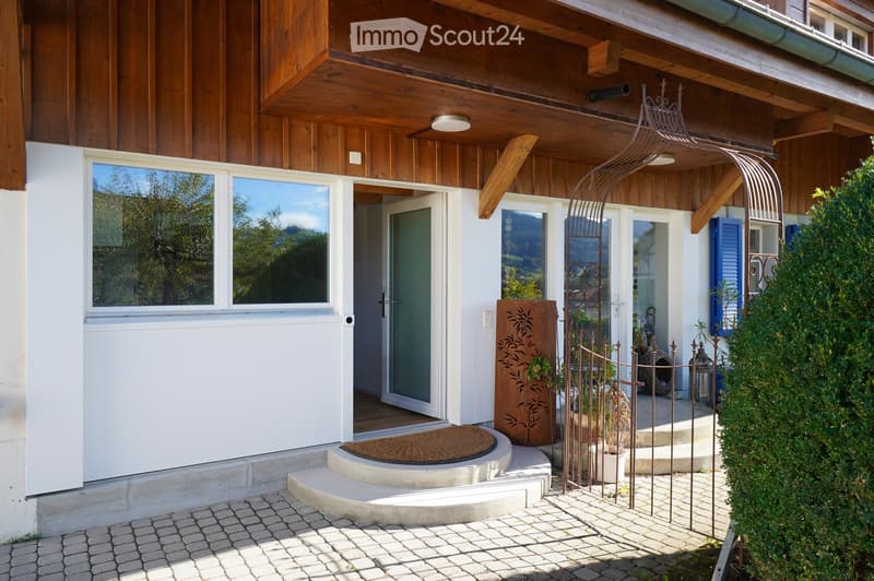 Front entrance of a two-story house with wooden siding, large windows, and a decorative metal gate leading to a small garden area.