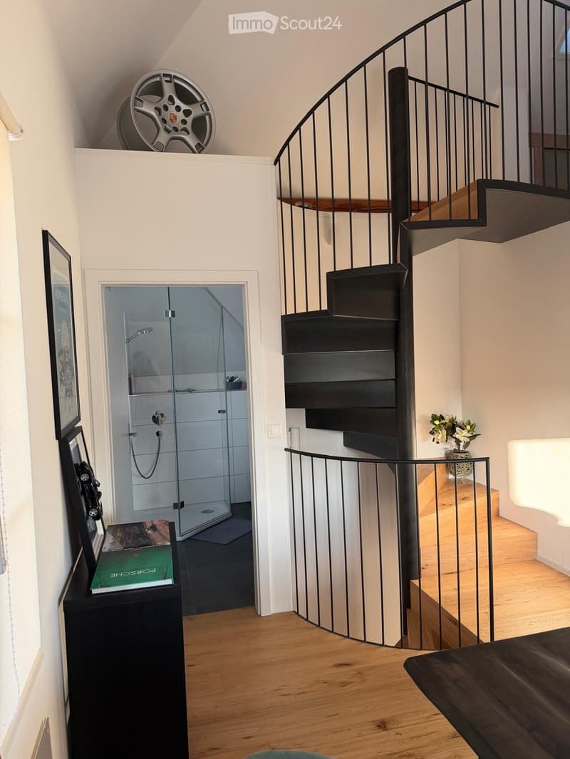 Spacious hallway with wooden flooring, modern staircase with black metal railing, glass-enclosed shower area, small table with vase of flowers, and framed artwork on the wall.