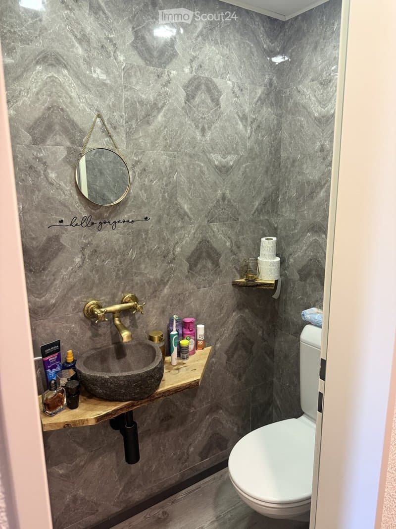 Modern bathroom with marble walls, a stone sink, brass faucet, a round mirror, and a wooden shelf holding toiletries and a toilet paper roll.